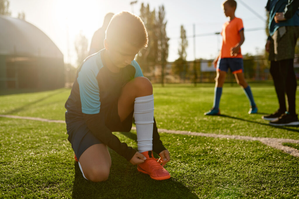 A young soccer player kneeling on the field to tie his cleats before practice, teammates in the background.