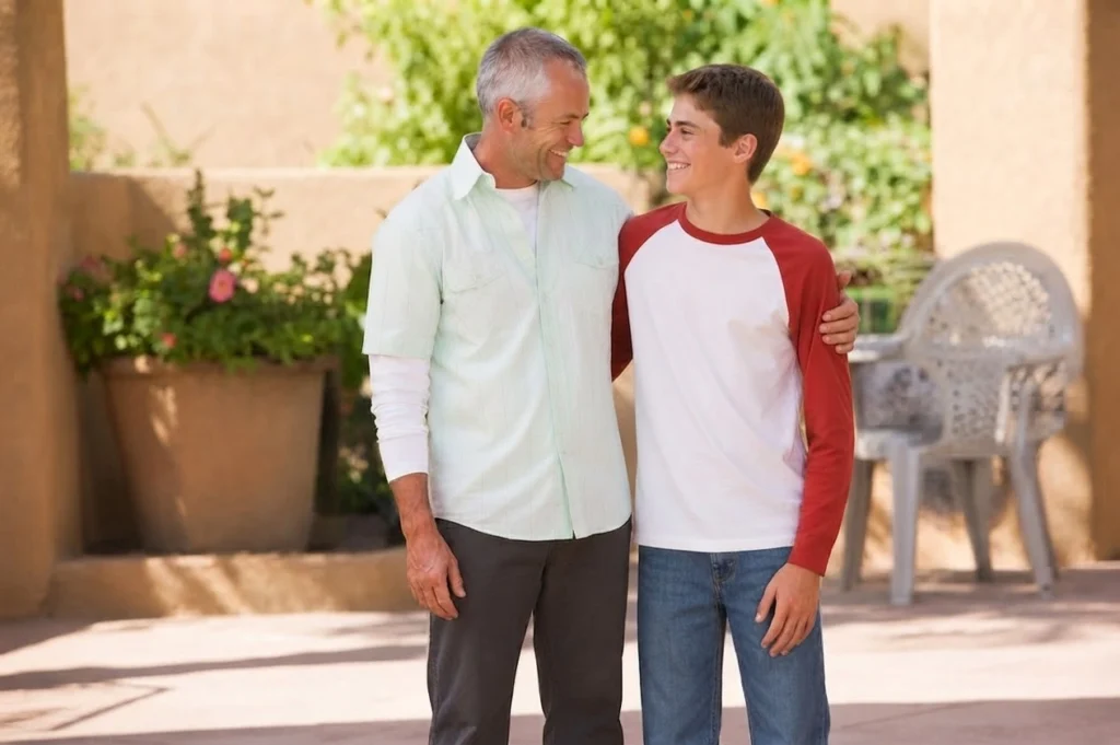 A father with his arm around his teenage son, smiling together outdoors.