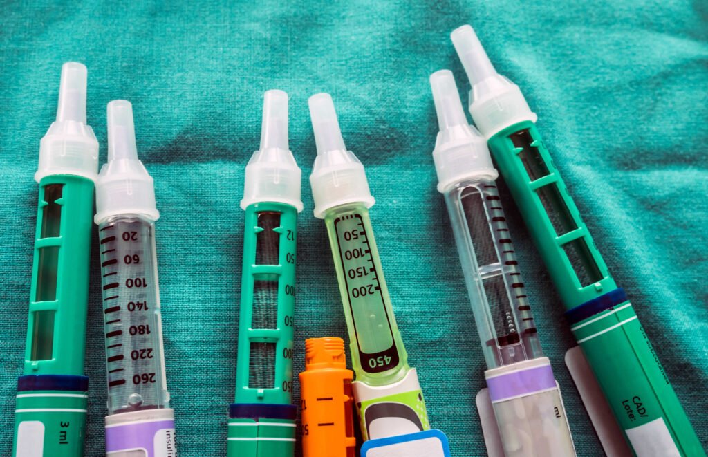 Several hGH injection pens with different sizes and dosing scales laid out on a teal surface, illustrating that growth hormone brands come in different delivery devices despite containing the same active hormone.