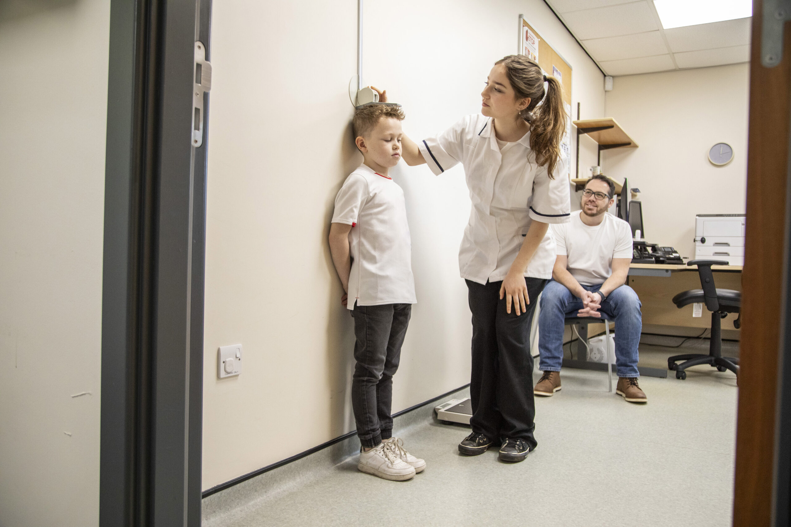 Nurse measuring child height at clinic wall chart during pediatric growth checkup with parent present