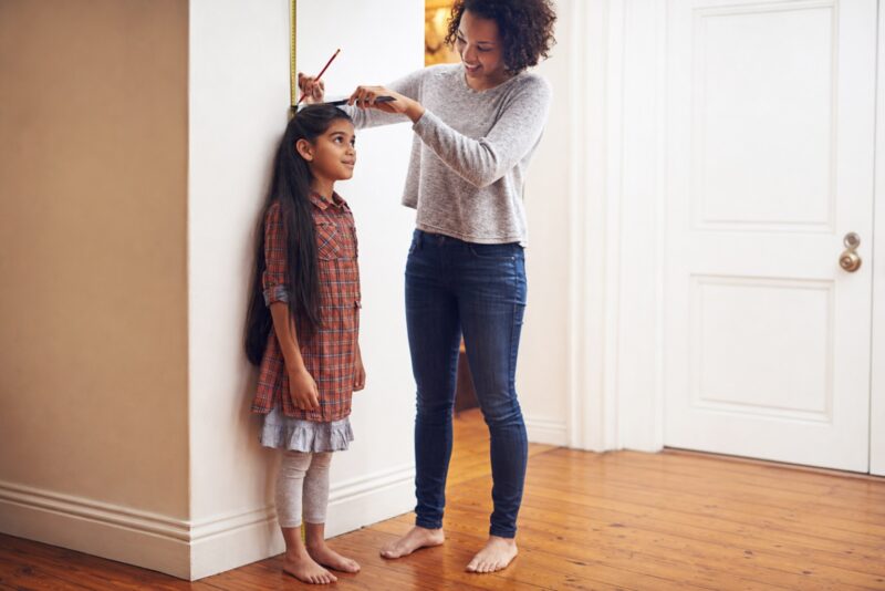 Mother measuring daughter's height against a wall at home, tracking child growth